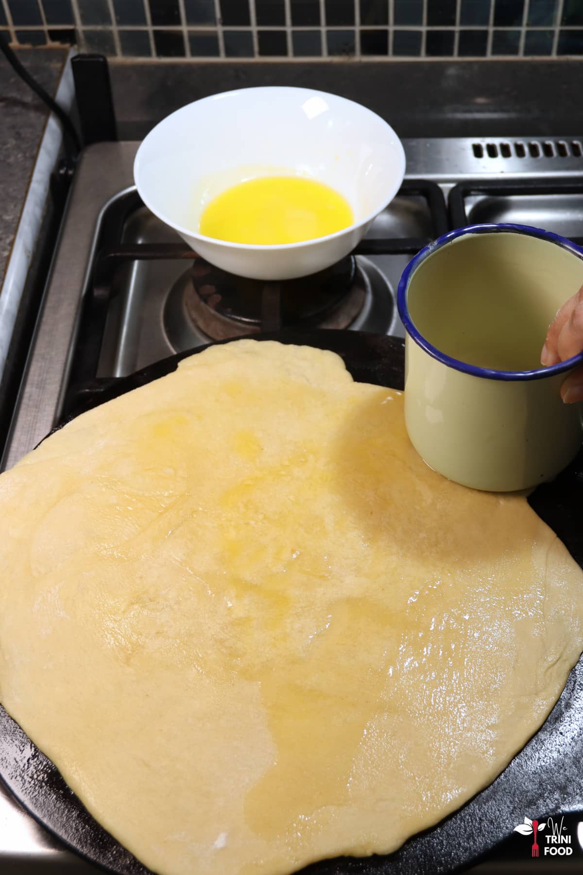 add oil or butter to buss up shut roti while it is cooking on a tawa with a bowl of butter on the side and an enamel cup being used to spread the butter