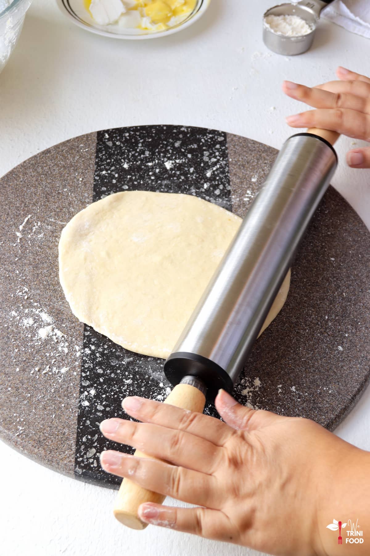 rolling out roti with rolling pin on a gray and black surface with butter and shortening on a plate in the background and flour in a measuring cup