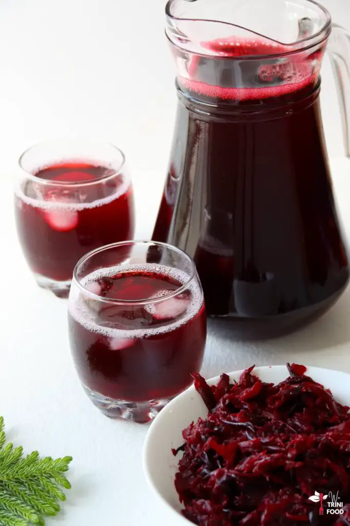 2 glasses of sorrel drink with ice and a jug of sorrel with a small christmas tree leaf and strained sorrel bits in a white bowl