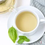 basil mint tea in a white teacup with basil and mint leaves on a saucer with tea remaining in a tea infuser