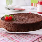 trinidad black cake or fruit cake garnished with cherries and mint leaves with a cake cutter and bottle of cherries in the background and a red striped tea towel in the foreground
