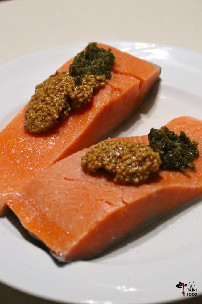 2 raw salmon fillets on a white plate with wholegrain mustard with honey, oil, salt and green seasoning