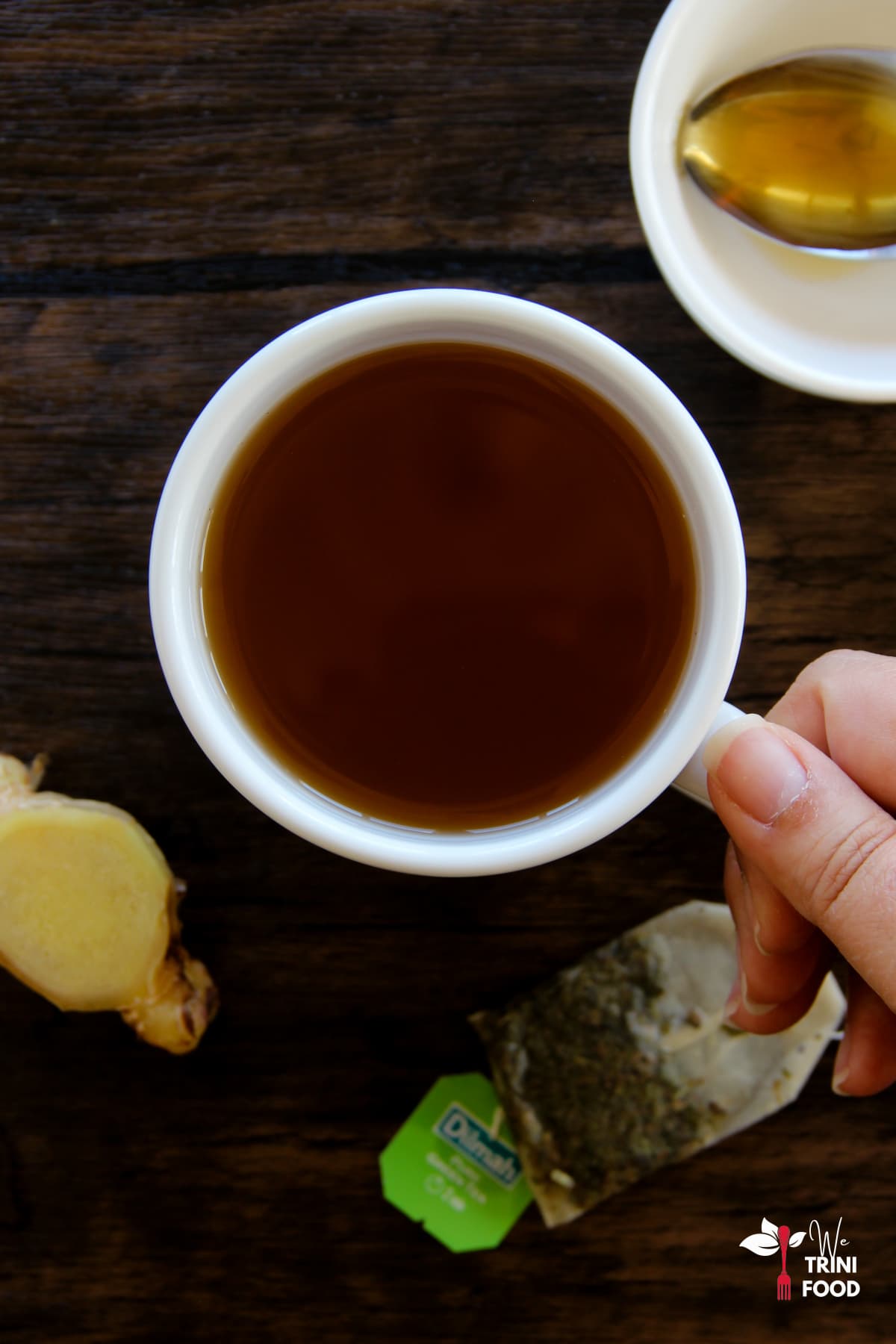 holding a cup of green tea with ginger with honey in a teaspoon, ginger and green teabag around the teacup