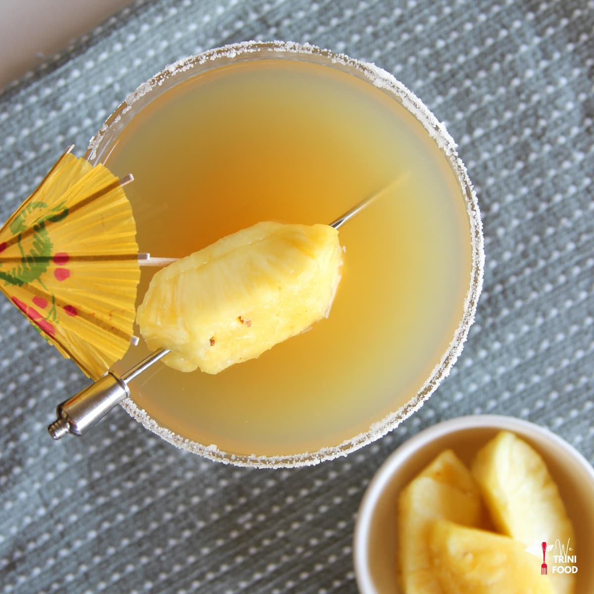 pineapple margarita cocktail in a margarita glass with pineapple and cocktail umbrella as garnish with a small bowl of pineapple wedges on the side
